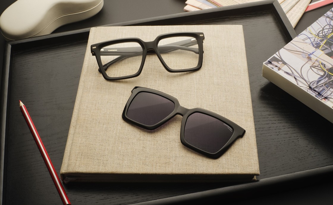 Carrera: Schwarze Sonnenbrille und schwarze Brille liegen auf einem Schreibtisch